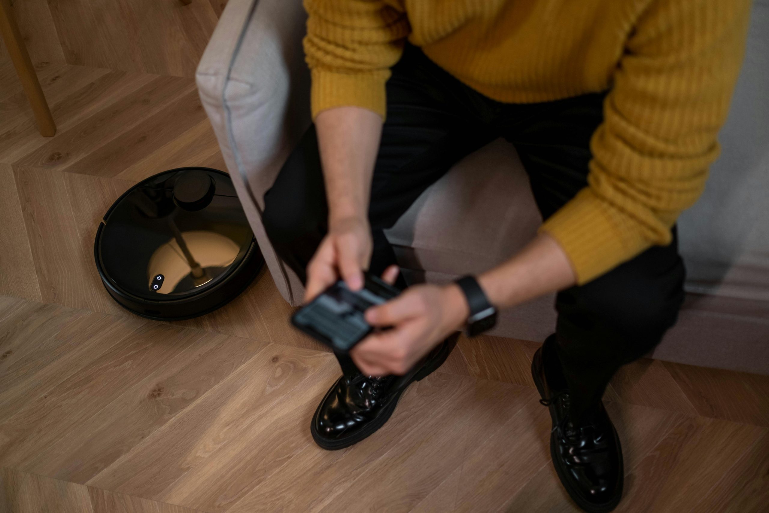 Man in yellow sweater using remote to control robotic vacuum on wooden floor — illustrating: Which robot vacuum actually handles dog hair without breaking the bank?