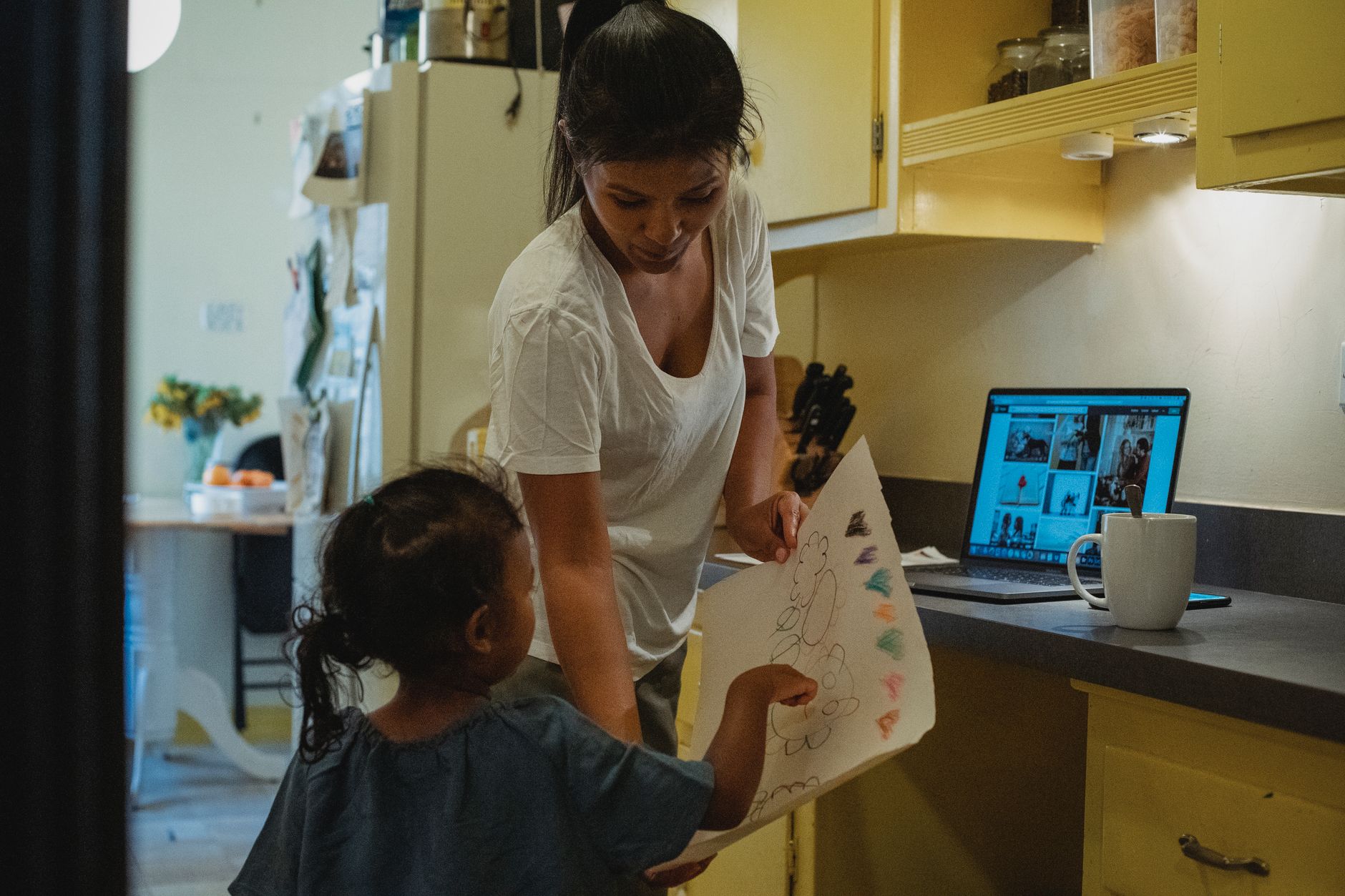 Unrecognizable little ethnic girl demonstrating paper sheet with drawing to mother while standing in kitchen with laptop and cup of drink — illustrating: Photo editing software for parents: what the reviews say about which tools are actually worth your time